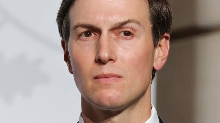 A close-up portrait of a man with short brown hair and a serious expression, wearing a suit and tie.