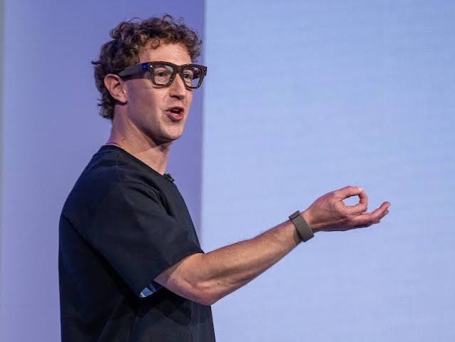 A person speaking on stage, wearing glasses and a black shirt, gesturing with one hand.