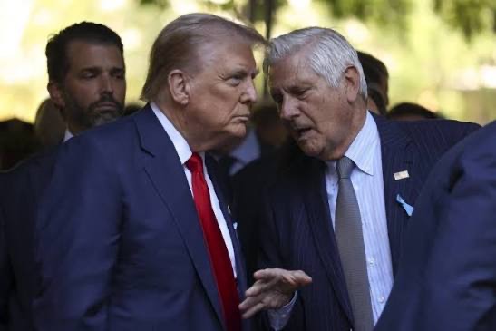 Former President Donald Trump engaged in a conversation with another man, with a serious expression. A person is partially visible in the background.