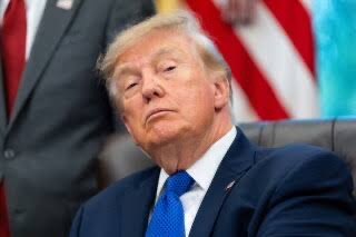 A close-up of a man with blonde hair wearing a dark suit and blue tie, seated and looking pensively to the side, with American flags in the background.