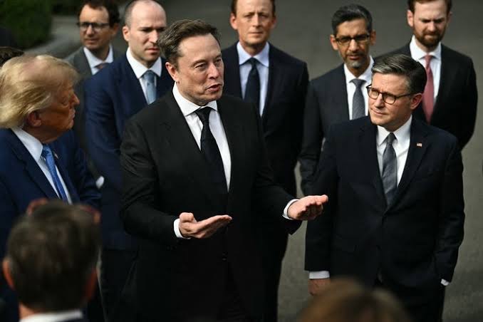 Elon Musk speaking to a group of individuals, including Donald Trump, during an outdoor event.